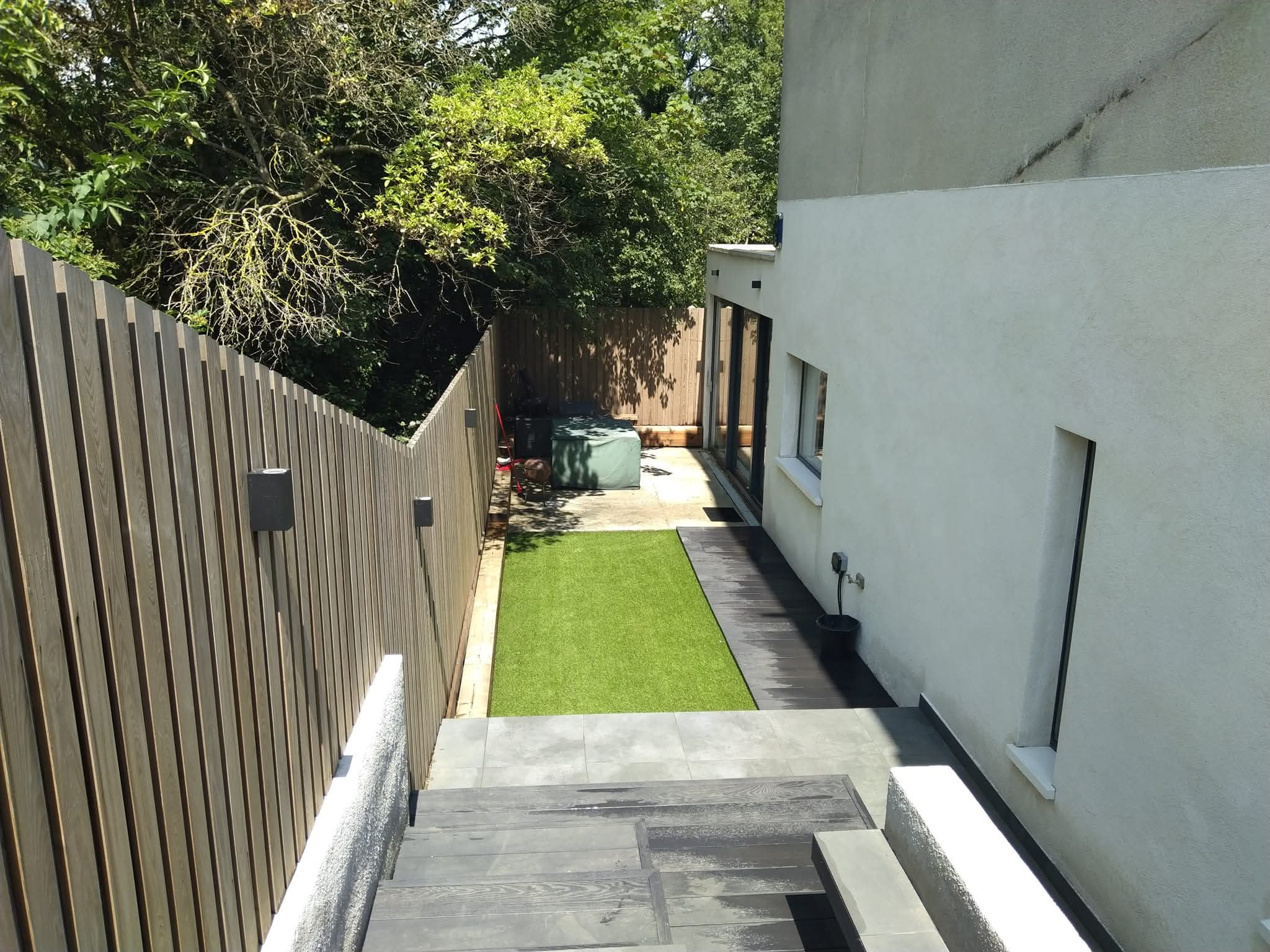 Haya UK gallery photograph of a narrow garden with green lawn, stone paving, steps and timber fencing.